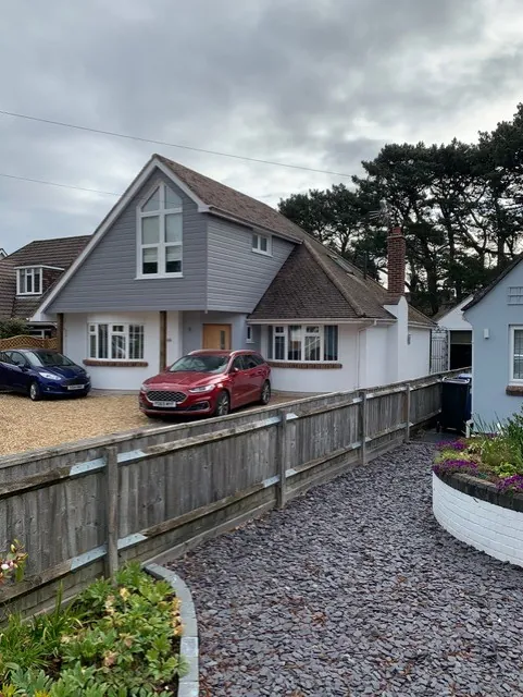Front of the finished house, with a grey clad gable and tall gable window above a white rendered ground floor, and a gravel drive