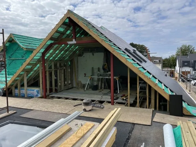 A gable end loft conversion part built, with red steel posts and a beam inside, green breathable membrane over the rafters and the first roof tiles laid on one side