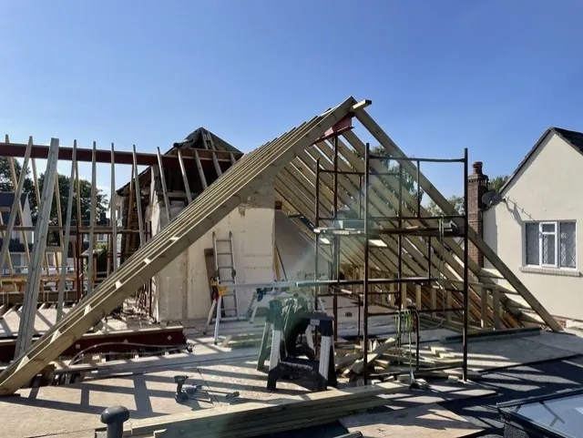 Timber rafters of a new gable end being built around red steelwork, with a scaffold tower and a mitre saw on the roof deck