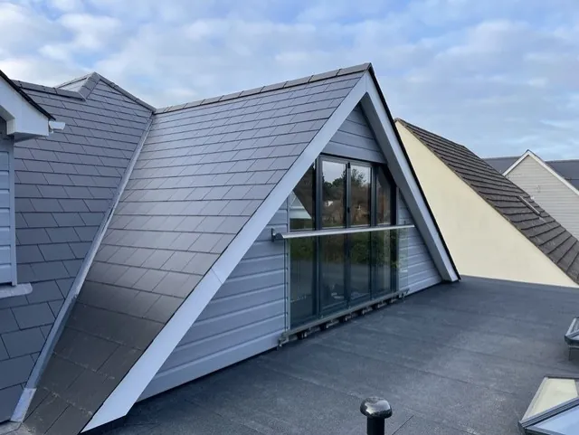 Finished gable end clad in grey weatherboard, with full height glazing and a glass balustrade, rising from the extension’s flat roof